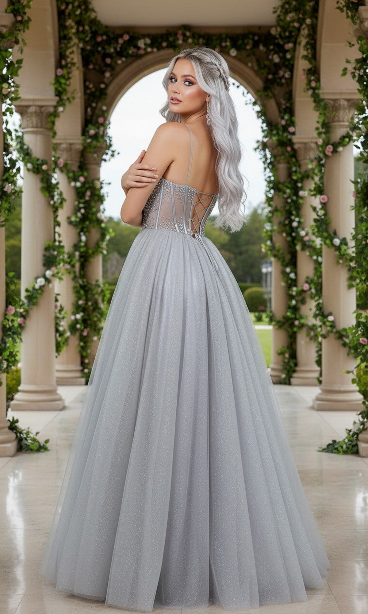 A woman with long silver hair wears the Cinderella Couture 8459J long prom dress, featuring a light gray, floor-length A-line tulle skirt, as she stands in an outdoor archway adorned with greenery and flowers.